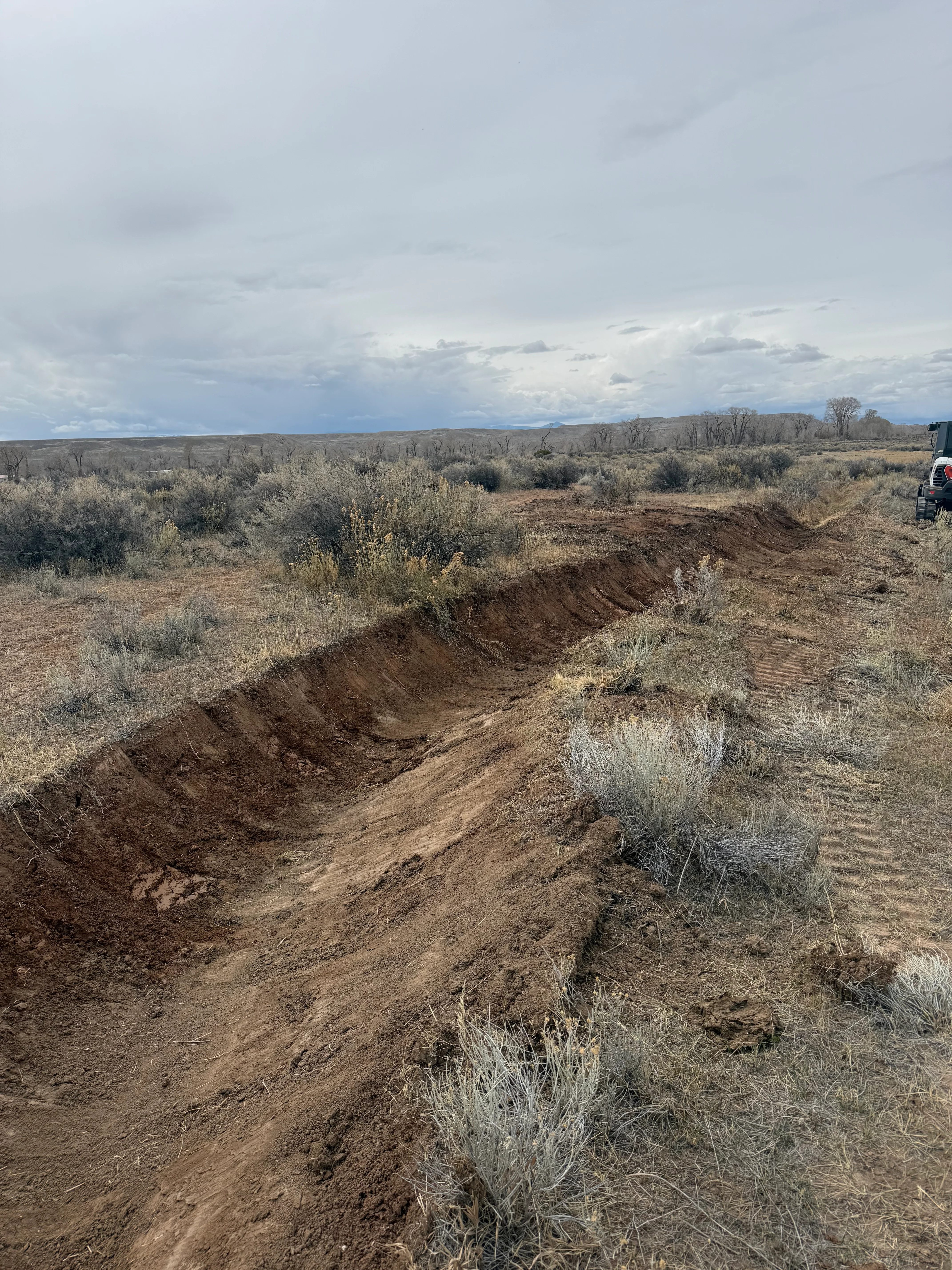 Graded earth trench and excavated soil on sagebrush rangeland with equipment visible