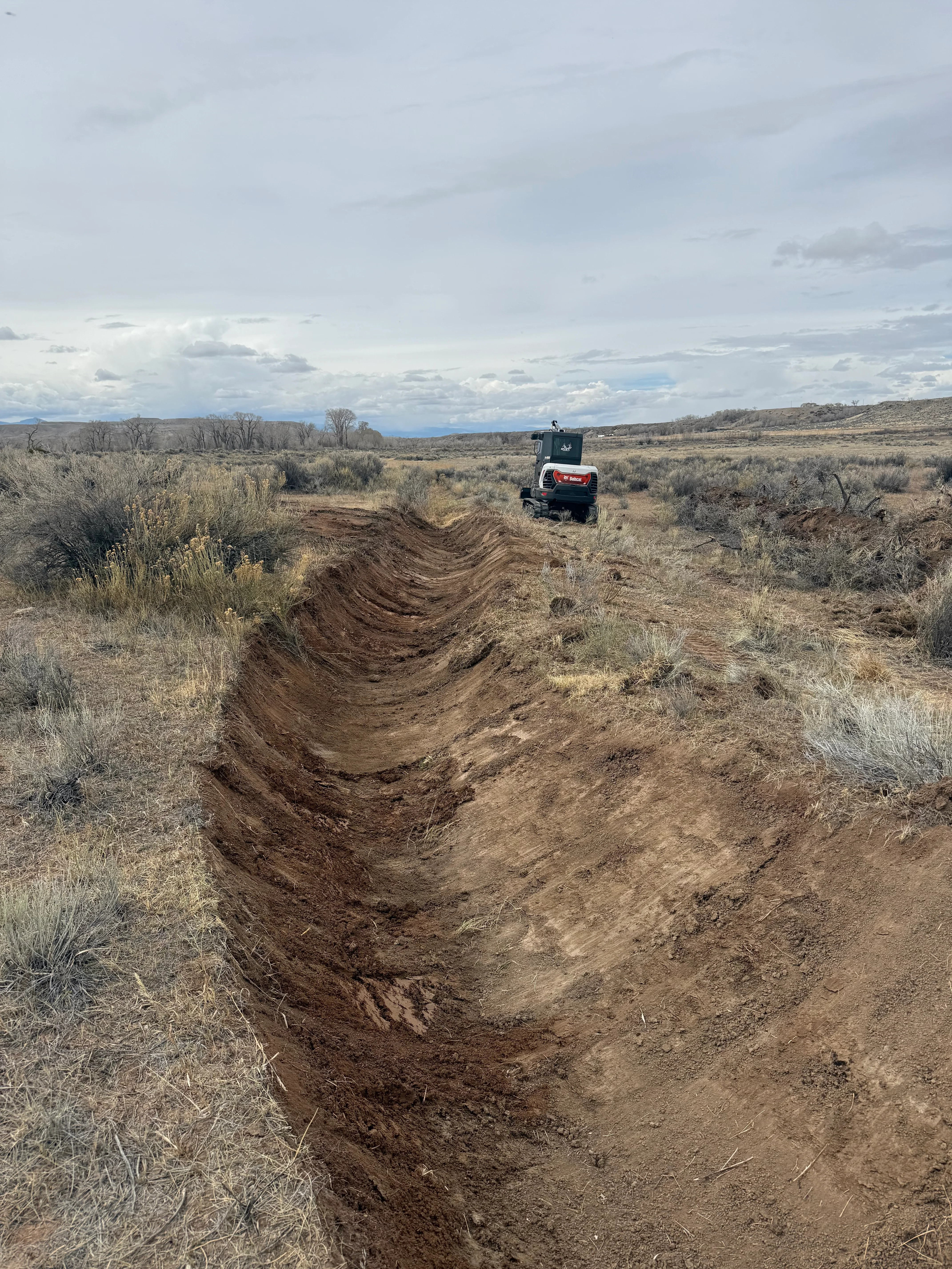 Tracked excavator creating deep drainage trench through sagebrush terrain with clean soil walls