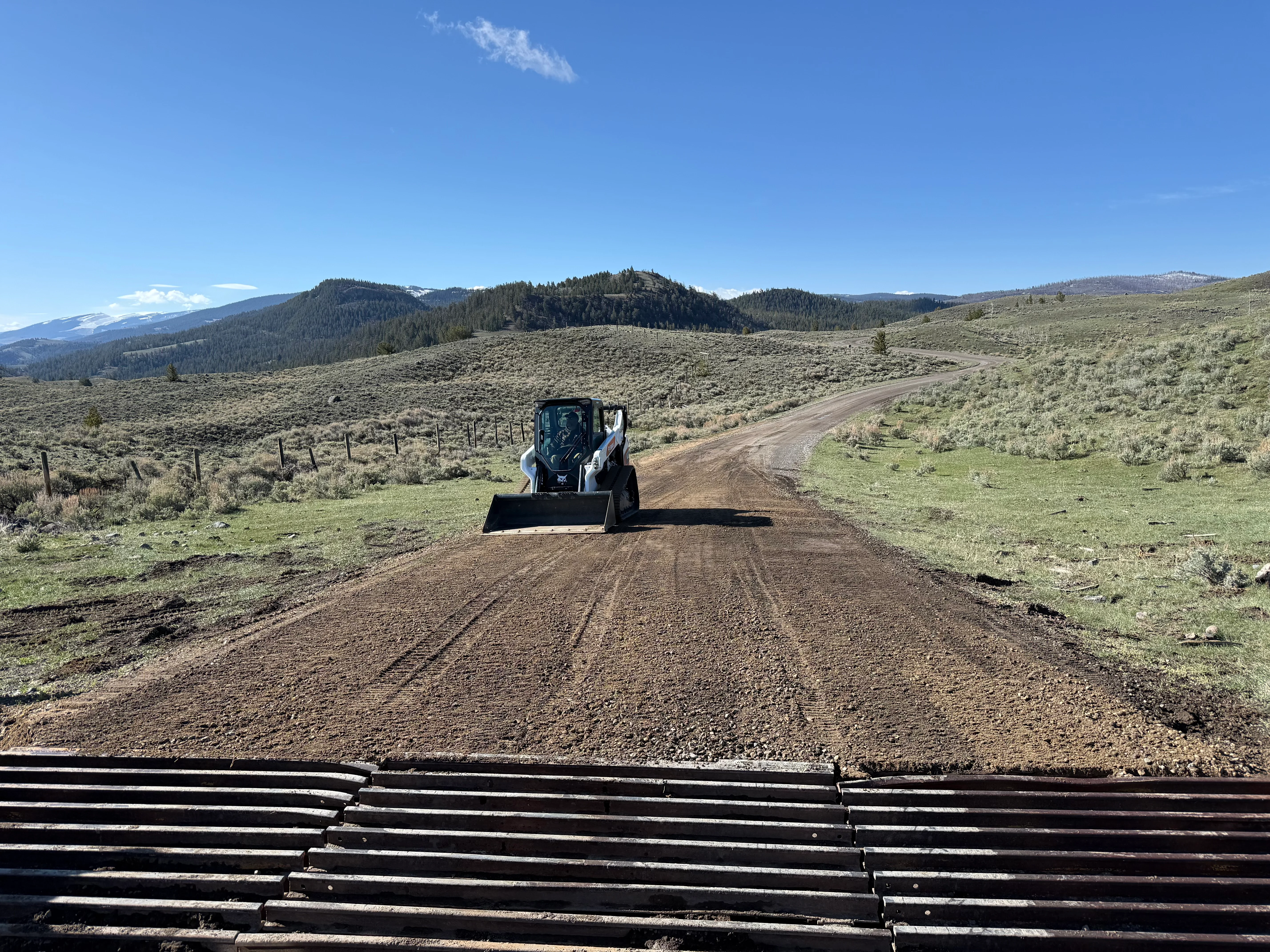 Skid steer loader grading dirt driveway through sagebrush terrain with mountains in background