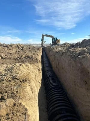 Excavator installing large corrugated black drainage pipe in deep trench with earthwork