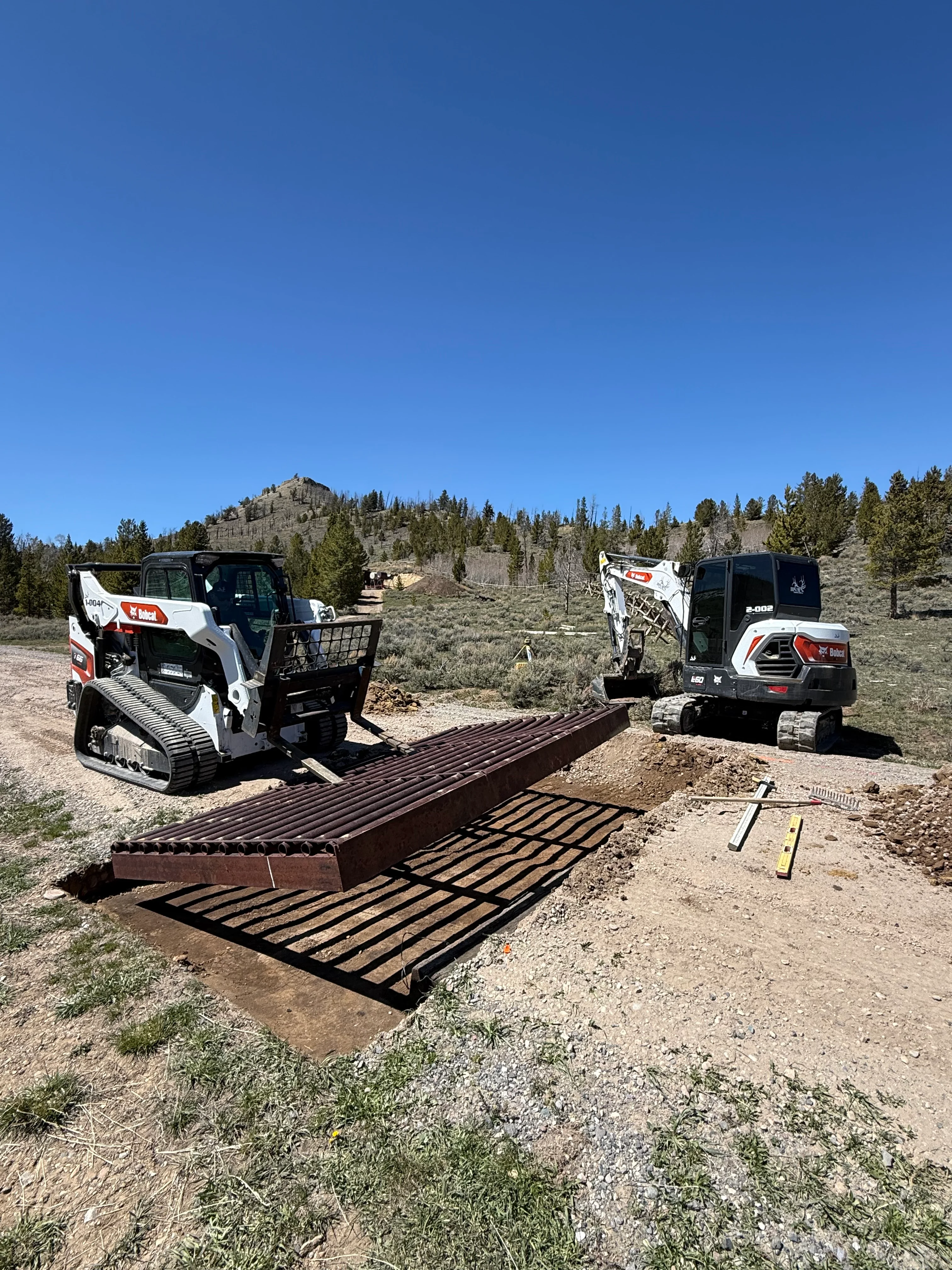 Two compact track loaders installing metal gate sections on gravel base in rural setting